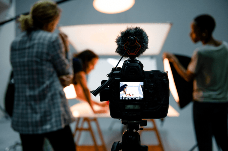 A film camera is set up, with people moving lights and objects in the background