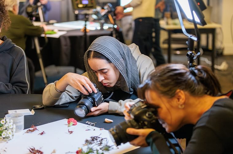 A group of people are holding cameras, aiming them at a black table that is set up with objects to photograph