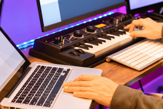 A person is at a desk, playing the mini keyboard with one hand, their other is placed on a laptop