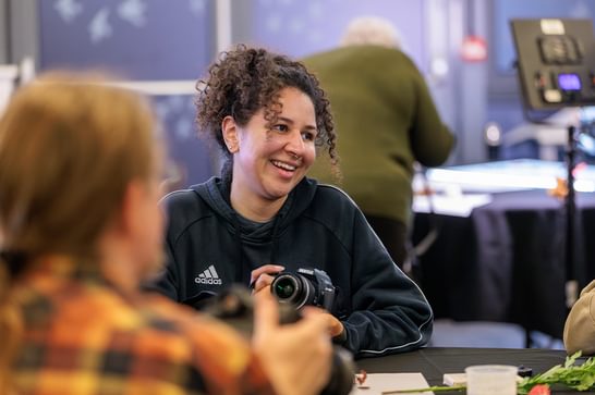 A woman is sitting around a table, holding a camera and laughing to the person next to her