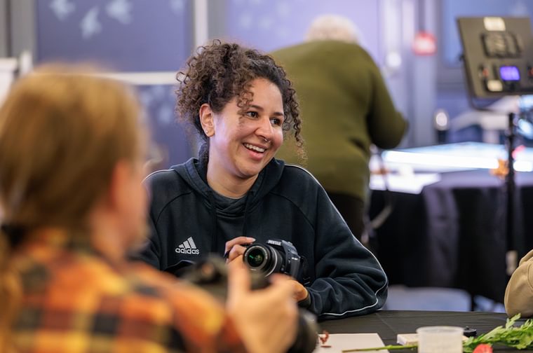 A woman is sitting around a table, holding a camera and laughing to the person next to her