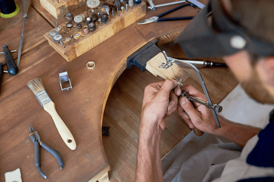 A person at a jewellery bench, sawing a metal ring on a table surrounded by tools