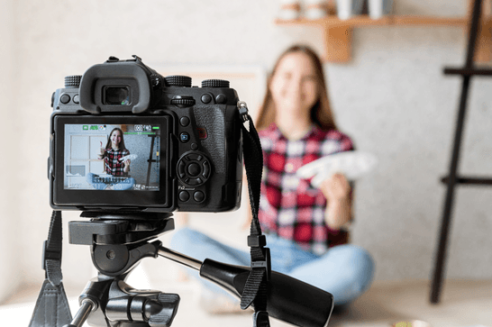 A black camera to the left, with a blurred background. On the screen of the camera, a teenage girl is smiling and putting her thumb up