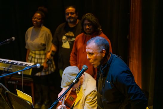 A group of people gather on stage, to the left a man sings into a microphone