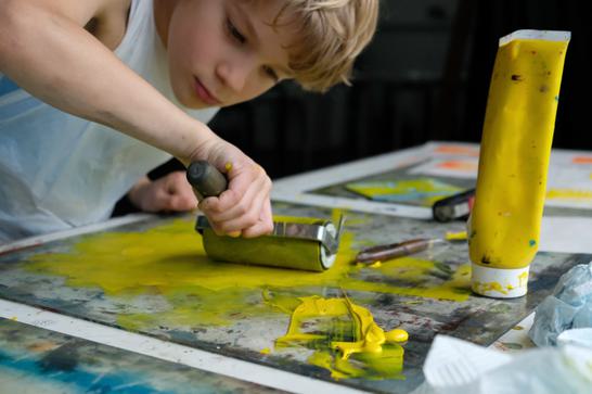 A young child is rolling out yellow ink on a transparent board, ready to print with