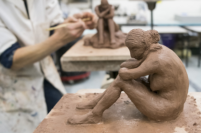 A nude human figure, sat with their elbows on their bent knees is made out of clay