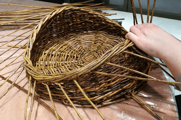 A person is weaving wooden willow sticks into a basket with a wavy top