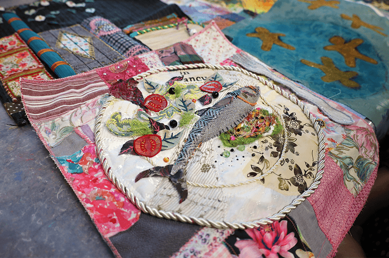 An embroidery piece of a fish a salad on a plate, surrounded by pink patch fabric