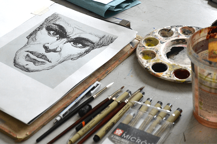 A table with a face drawing, pens, paint brushes and a palette on it