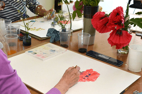 People are sat at a table, with a selection of flowers. They are painting in watercolours, using the flowers in front of them as inspiration.