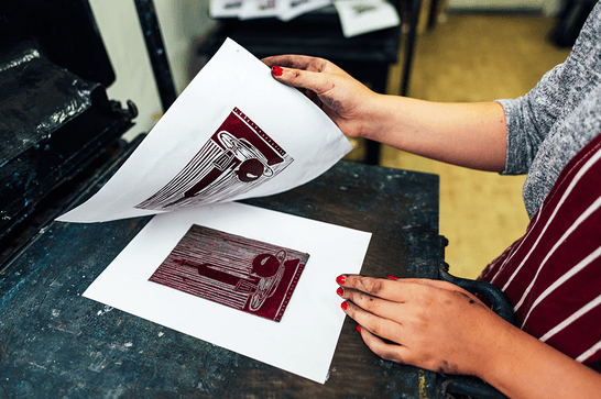A person is lifting a piece of paper off a cut lino block, revealing a print of still life objects in mahogany ink