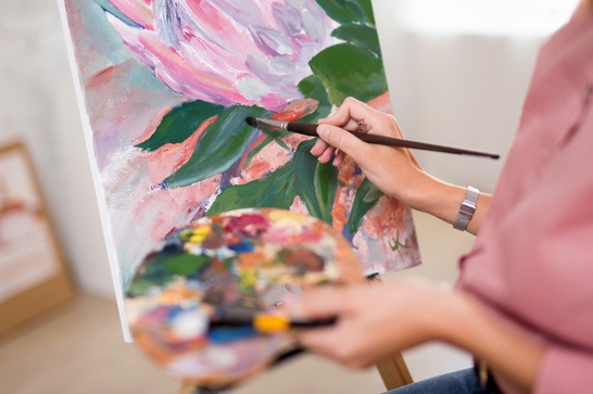 A person is holding a paint pallet and brush, painting green leaves and pink flowers on a canvas in front of them