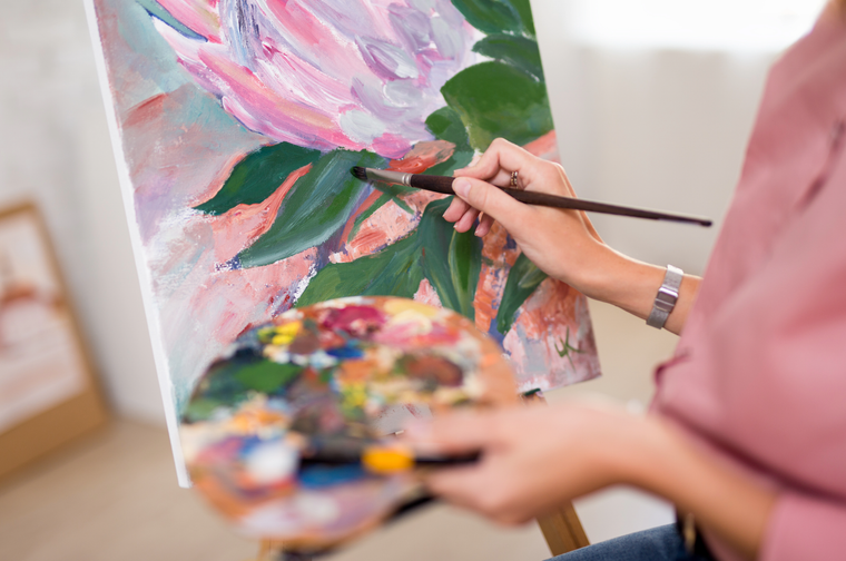 A person is holding a paint pallet and brush, painting green leaves and pink flowers on a canvas in front of them