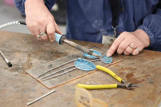 A person is using a soldering iron to connect a blue stained glass flower