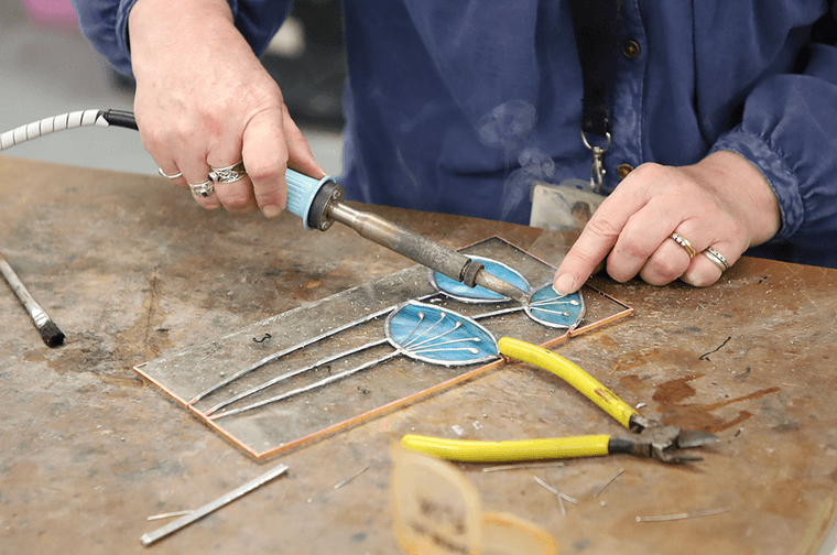 A person is using a soldering iron to connect a blue stained glass flower