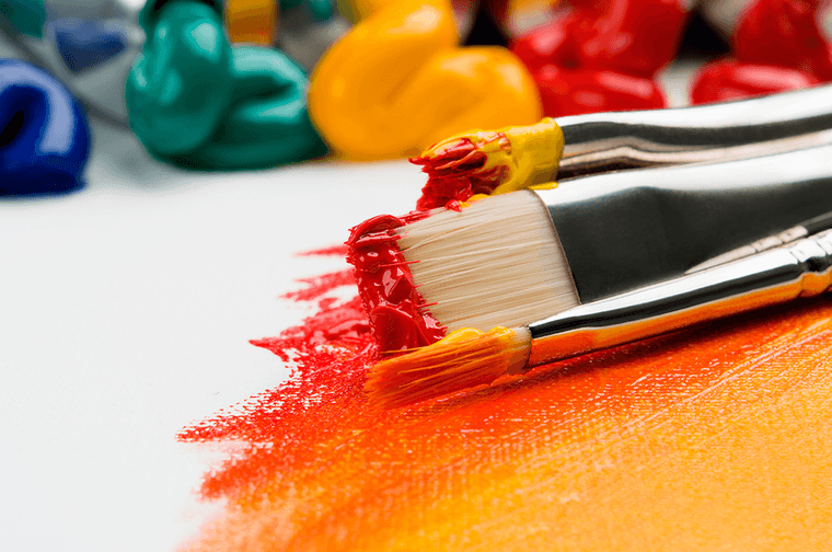 Paint brushes with yellow and red paint on them are resting on a canvas which is partially painted
