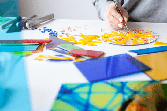A person is placing yellow glass into a pattern, at a table that has blue, green and yellow glass panels on it
