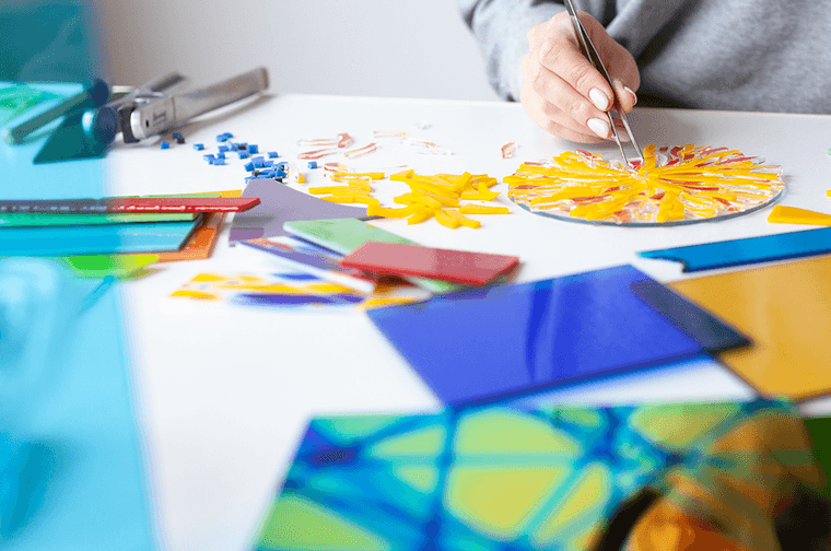 A person is placing yellow glass into a pattern, at a table that has blue, green and yellow glass panels on it