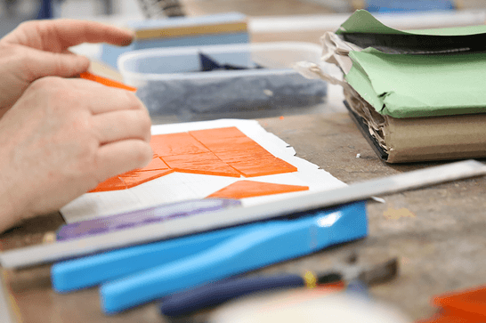 A person is arranging orange glass onto white paper, surrounded by tools
