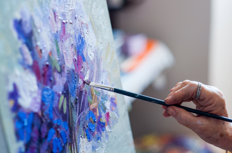 A person is painting a bunch of blue and purple flowers on a canvas with oil paints