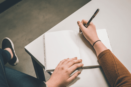 A person is writing in an open, blank notebook on a grey desk