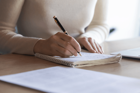A person is writing into an open notebook, with papers to the side of them with text on it