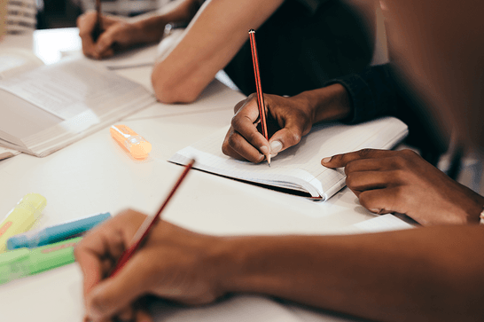 Three people are writing into open notebooks, surrounded by books and highlighters