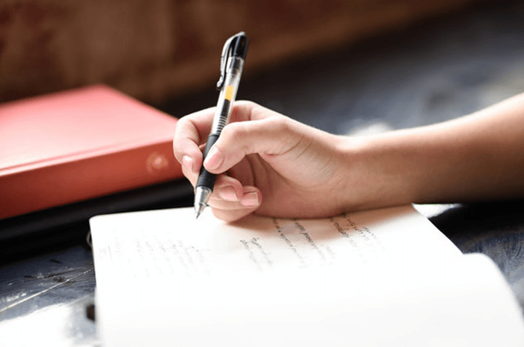 A person is writing in an open notepad, with books to the side of them