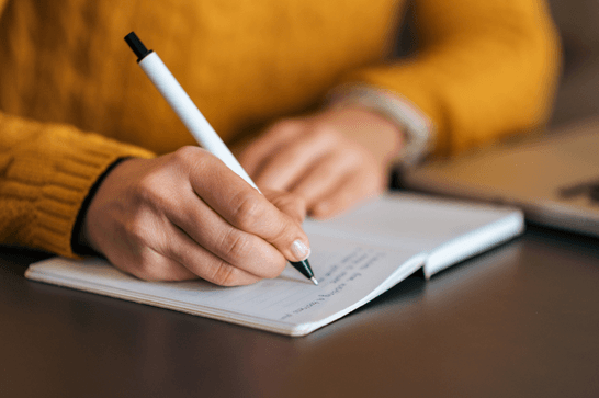 A person is writing in an open notepad and is wearing a bright yellow jumper