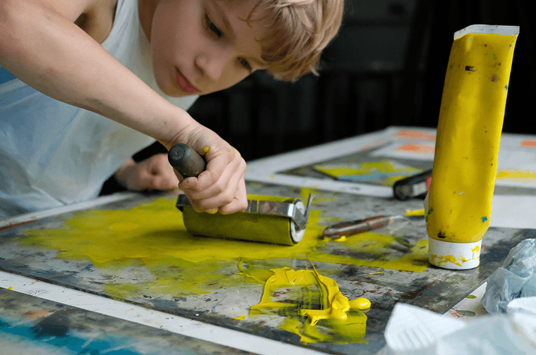 Young boy with blonde hair and wearing a white apron, making a bright yellow screen print.