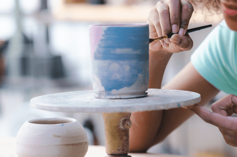 Close up of young girl painting a a blue and pink ceramic cup. The cup is on a wheel that she is holding with her left hand.