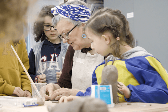 Young girl with braided hair and yellow and blue pinafore is playing with a block of clay. She is look at a woman on the left for guidence. The woman has shirt grey hair, a blue and white scarf on her head and is wearing a cream pinafore. She is making something out of clay on the table. There are two children behind her also watching her work.