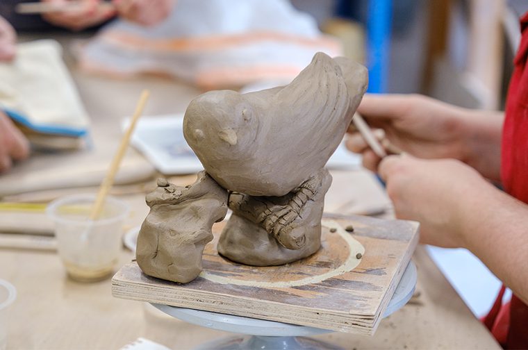 A small bird made from clay is on a wooden block, looking towards the viewer