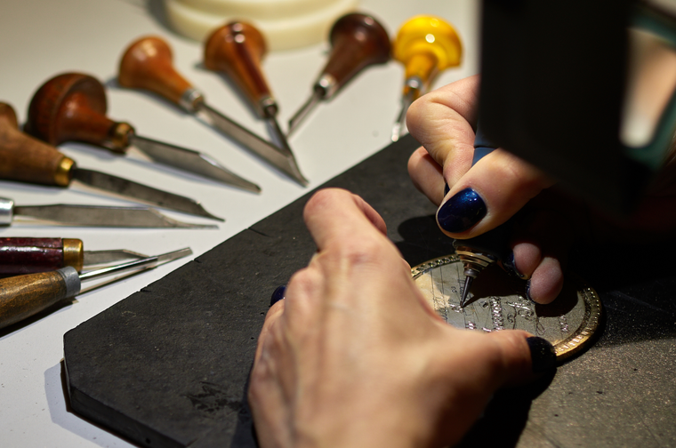 A person is engraving a small silver metal disk, with chisels surrounding the work area
