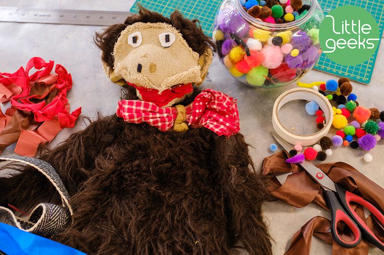 A toy monkey on a table, surrounded by crafting materials like pom poms and ribbons