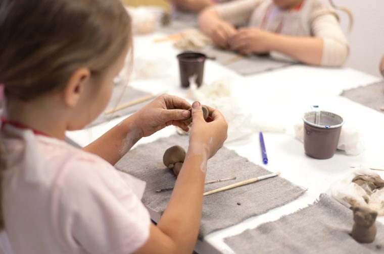Close up of young girl in a pink t shirt playing with clay. There is a mat on the table in front of her, some pottery tools and a cup of water.