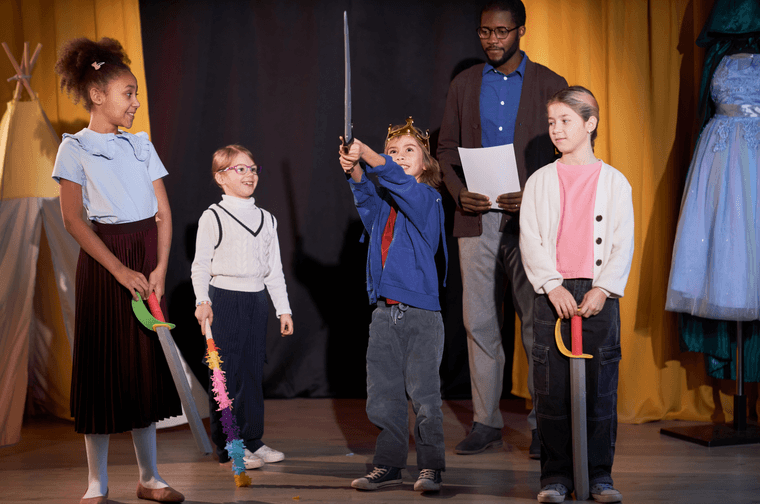 Group of children on a stage holding foam swords and acting. they are being directed by a man stood at the back of the stage with a script.