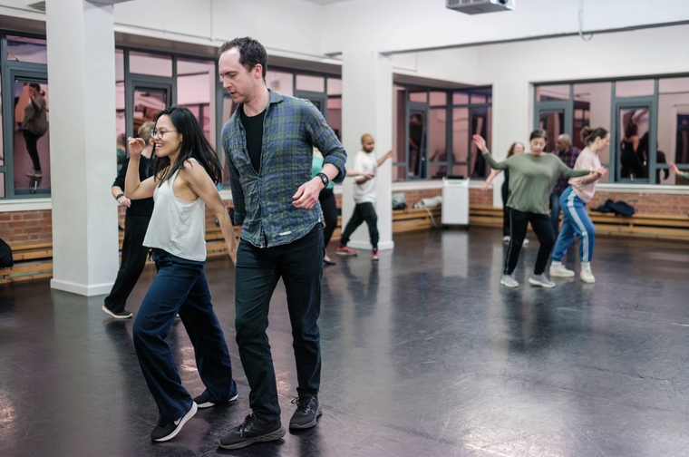 A group of couples is in a studio with a black floor, dancing the salsa