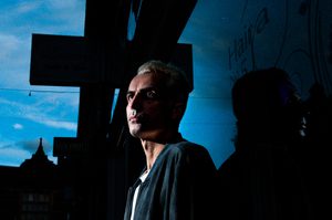 Dan is stood against a dark background, with dramatic shadows against Dan's face. Dan has short blond hair and is wearing a a navy shirt.