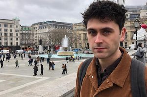Daniel is wearing a brown button up jacket and has short dark hair. Daniel is stood in front of a busy townscape.
