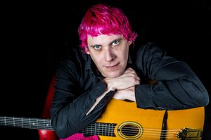 Gary is leaning onto his crossed arms, which are placed over a guitar. Gary has short pink hair.