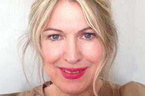 Hazel is standing against a white background, and is wearing a brown dress. Hazel has blond hair and is smiling.