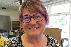 Helen is standing in our textile studio, and has short brown hair and a fringe. Helen is wearing a black and white floral blouse.