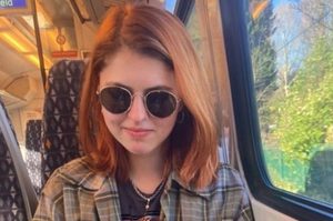 Georgia is seated on a train, and is wearing a checked shirt and sunglasses. Georgia has long, brown hair.