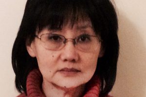 Mira is wearing a red jumper, has brown hair and a fringe and is wearing glasses.