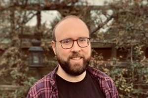 Neil Milton has a brown beard and is wearing a red checked shirt and jeans.