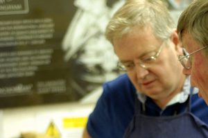 Nick is stood next to a table teaching. He has short grey hair and is wearing glasses.