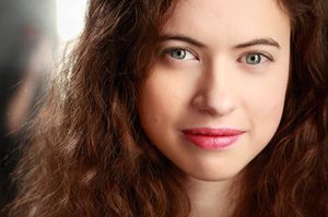 Roxanne has long brown curly hair and is looking directly to camera