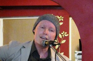 Tony is wearing a grey coat and grey hat and is sat at a microphone singing and playing the guitar.