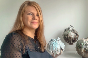 Andrea wears a black dotty top and long hair, and is next to exhibited ceramics